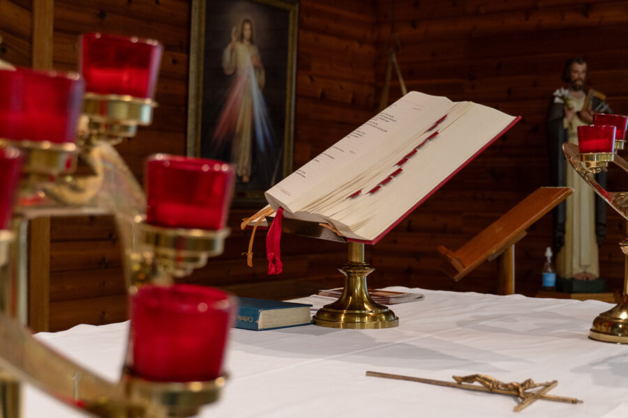 Catholic altar with lectionary and red votive candles in Our Lady of Fatima Church, Minden, Ontario – Open scripture book on brass stand with Divine Mercy and Saint Joseph in background.