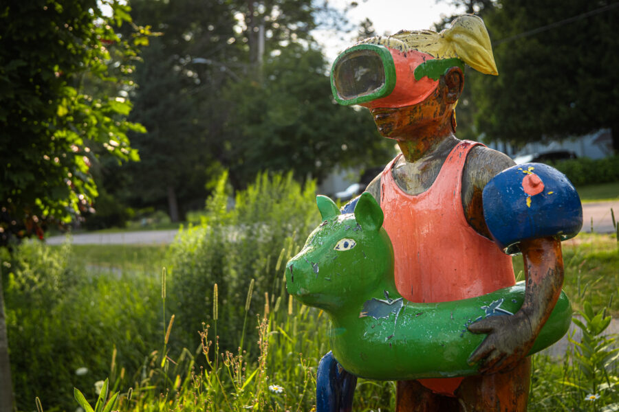 Wooden statue of a child in swim gear holding a green inflatable animal float, Minden Ontario